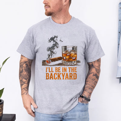 Black unisex backyard cigar and whiskey t-shirt with a lit cigar, bourbon on the rocks, and the text I'll Be in the Backyard