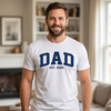 white unisex t-shirt with bold retro collegiate lettering reading DAD Est. 2026 in navy blue and white