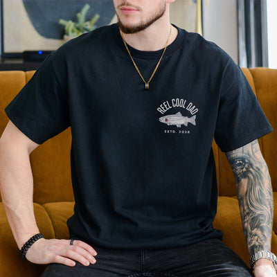 Ivory unisex t-shirt with a trout illustration and the words Reel Cool Dad plus a custom established year on the chest pocket area